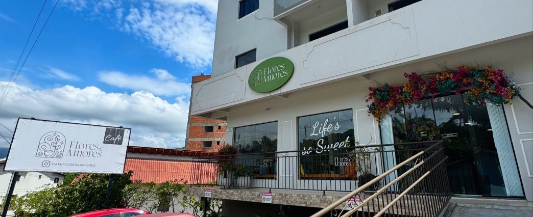Fachada do prédio branco com a placa verde do Flores e Amores, flores na porta e estacionamento na frente